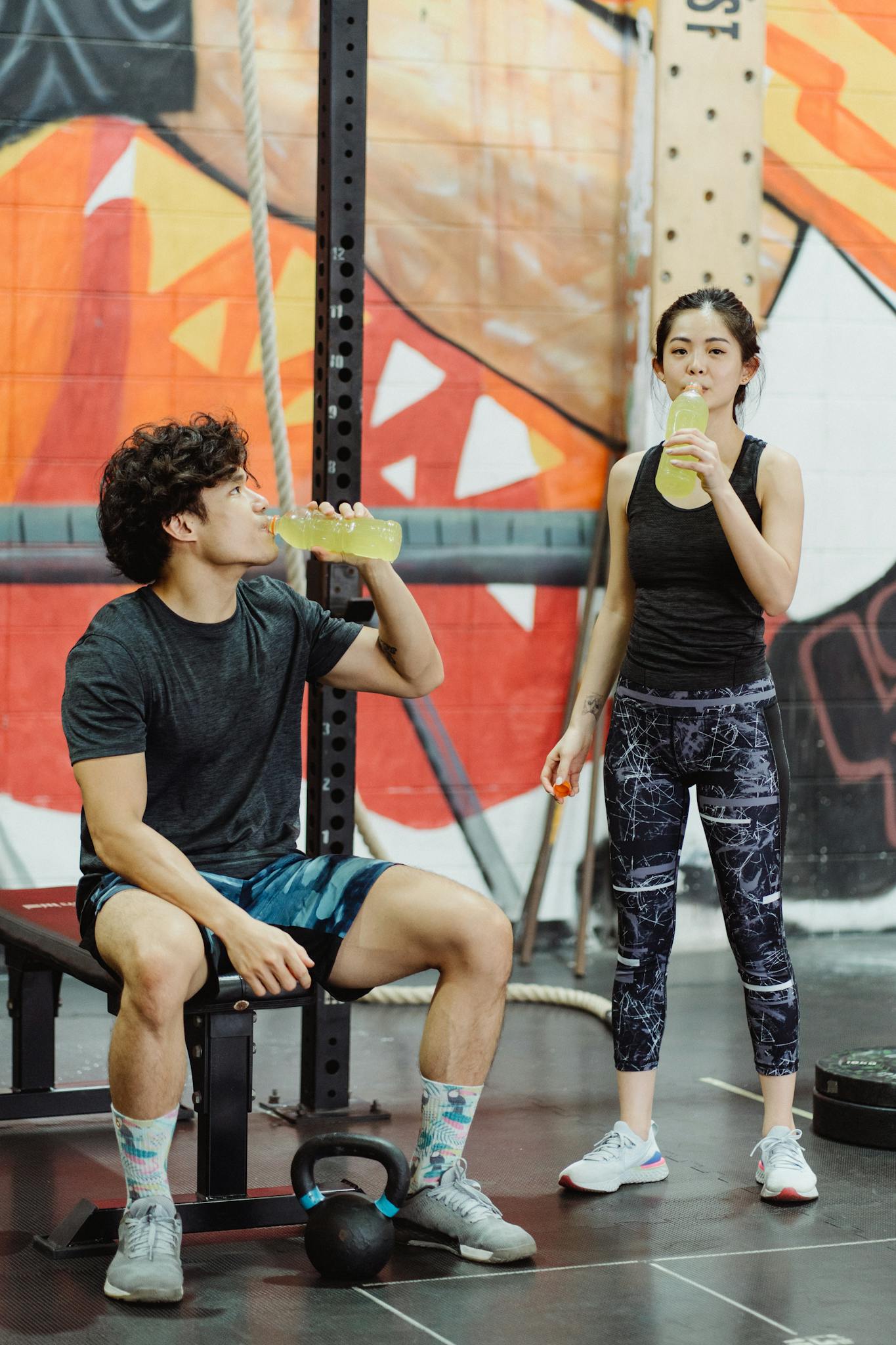 Asian man and woman in activewear drinking beverages at an indoor gym.